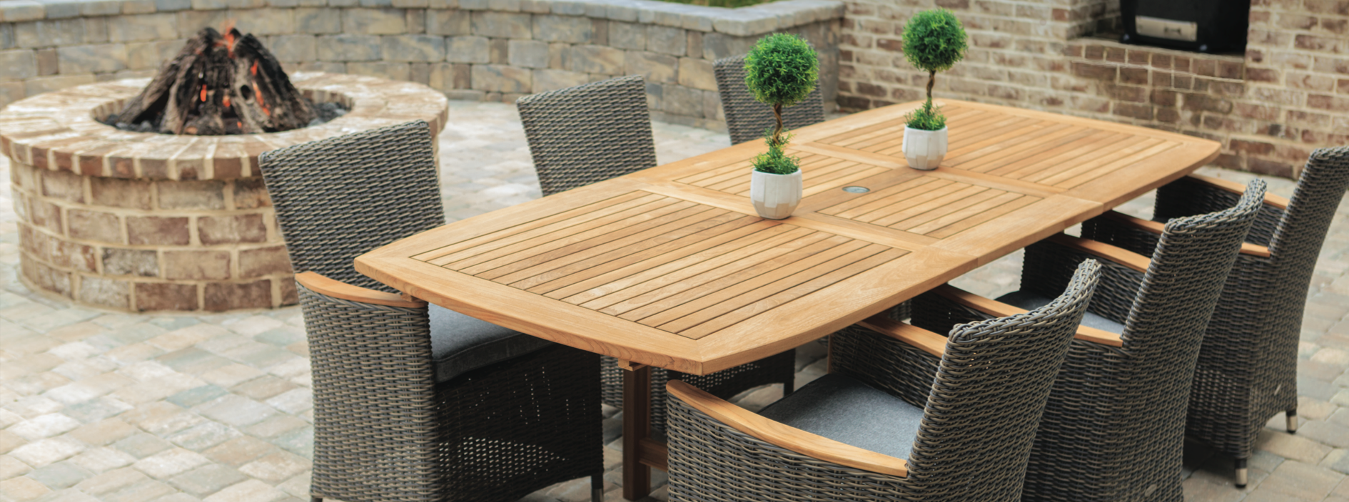 Patio with a teak table surrounded by 7 wicker chairs with an outdoor firepit in the background